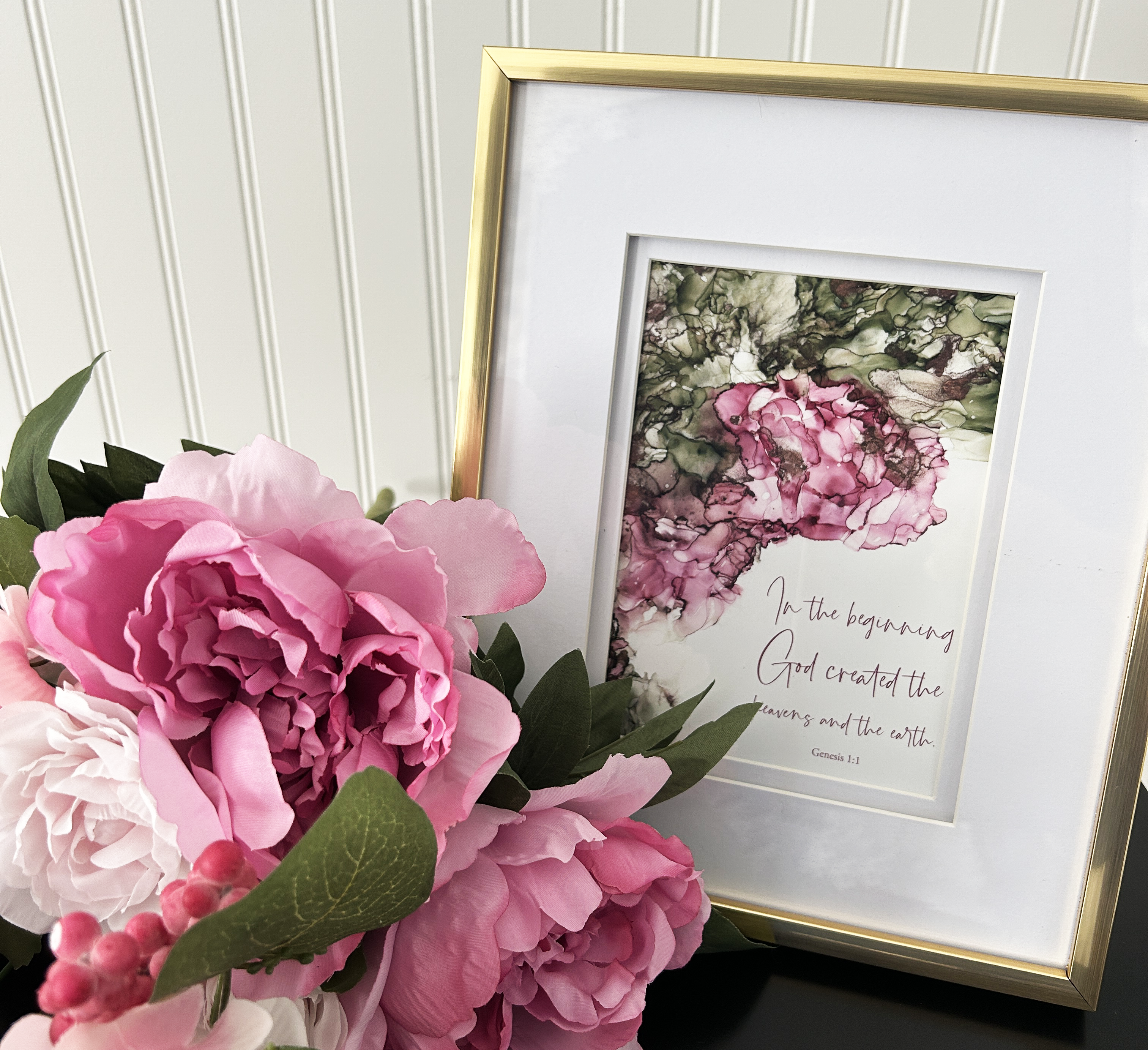 Framed Bible verse art print (Genesis 1:1) with pink flowers and greenery on a white wall background